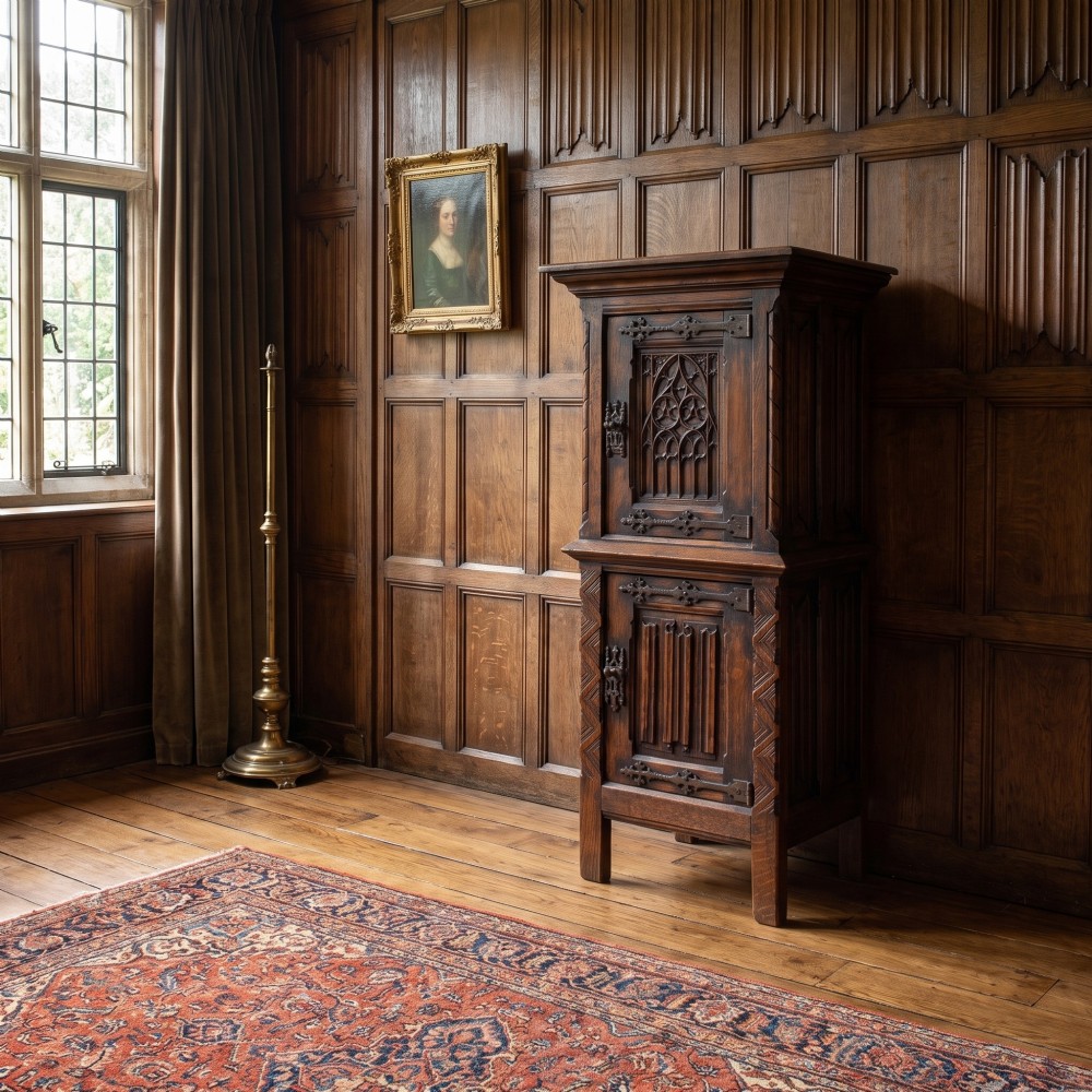 A tall, narrow dark oak Gothic Revival cabinet with two carved cupboard doors, decorative iron strap hinges, and a moulded top cornice. View II