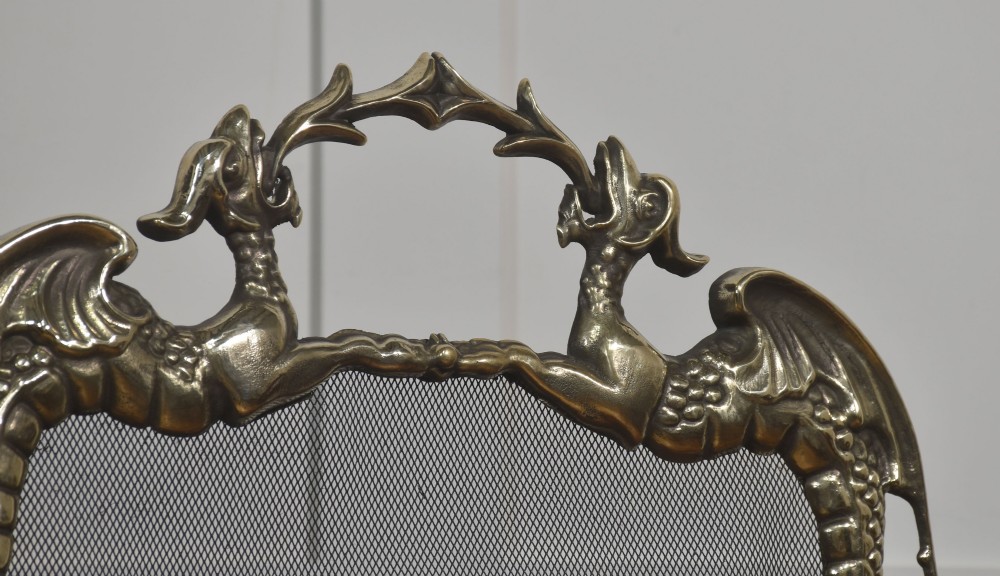 An ornate brass Rococo Revival fire screen featuring a central wire mesh panel, dragon-shaped top handles, and detailed claw feet. View V