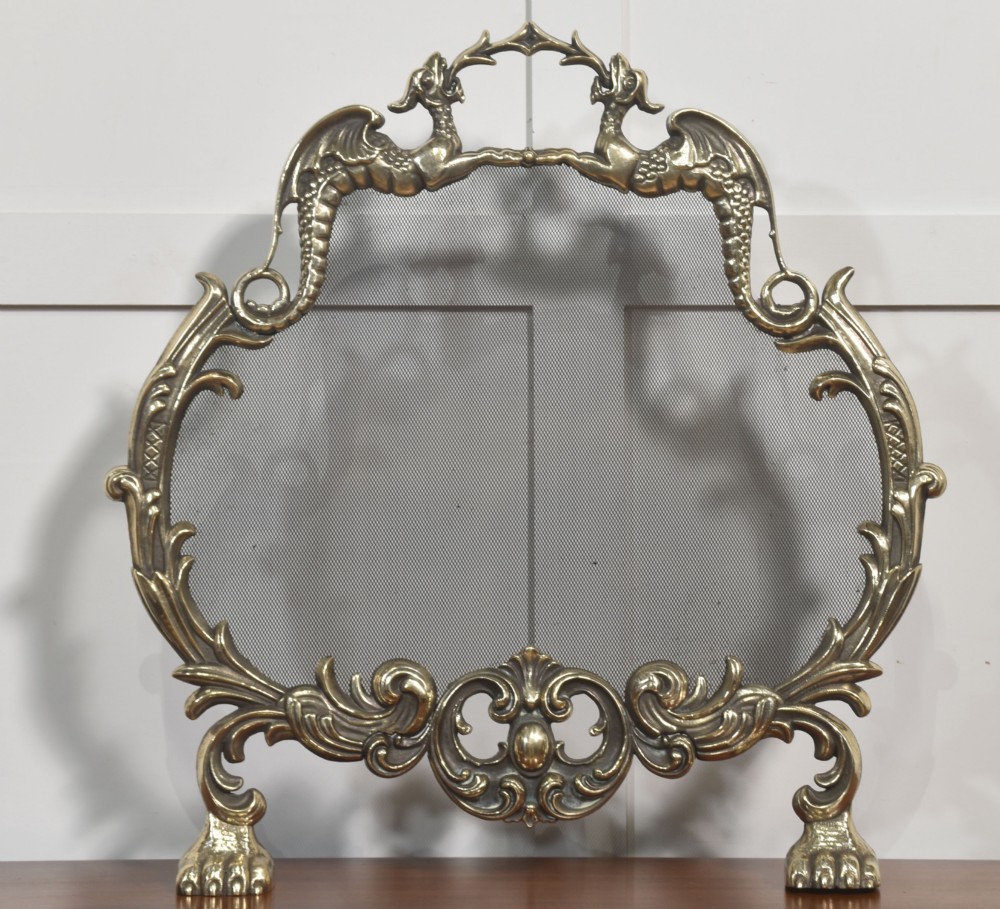An ornate brass Rococo Revival fire screen featuring a central wire mesh panel, dragon-shaped top handles, and detailed claw feet. View VI