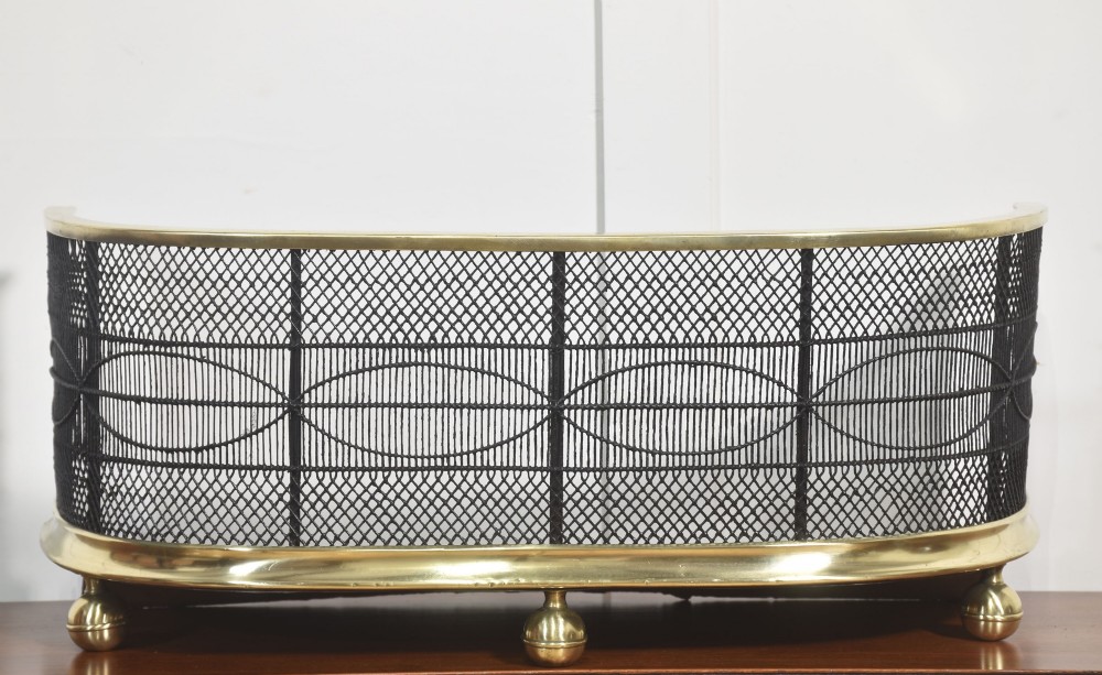 A curved antique fire guard made from black woven wirework with a polished brass top rim, base trim, and turned ball feet. View V