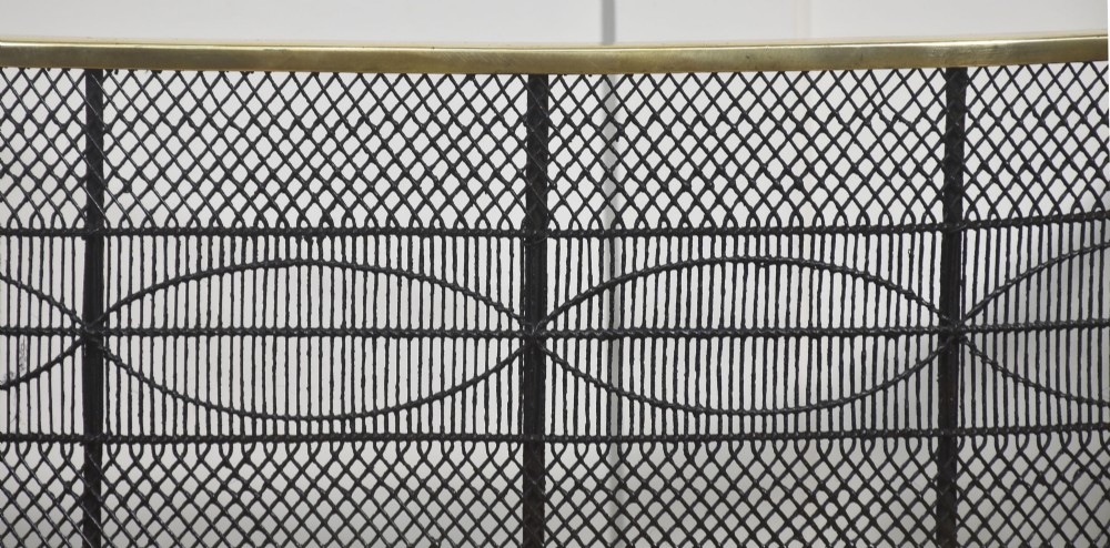 A curved antique fire guard made from black woven wirework with a polished brass top rim, base trim, and turned ball feet. View VI