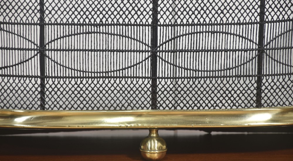 A curved antique fire guard made from black woven wirework with a polished brass top rim, base trim, and turned ball feet. View VII