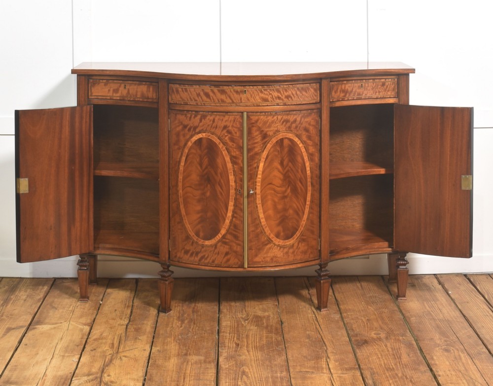 A Sheraton Revival bow-front wooden sideboard featuring oval inlaid veneer door panels, top drawers, and square tapered legs. View IX