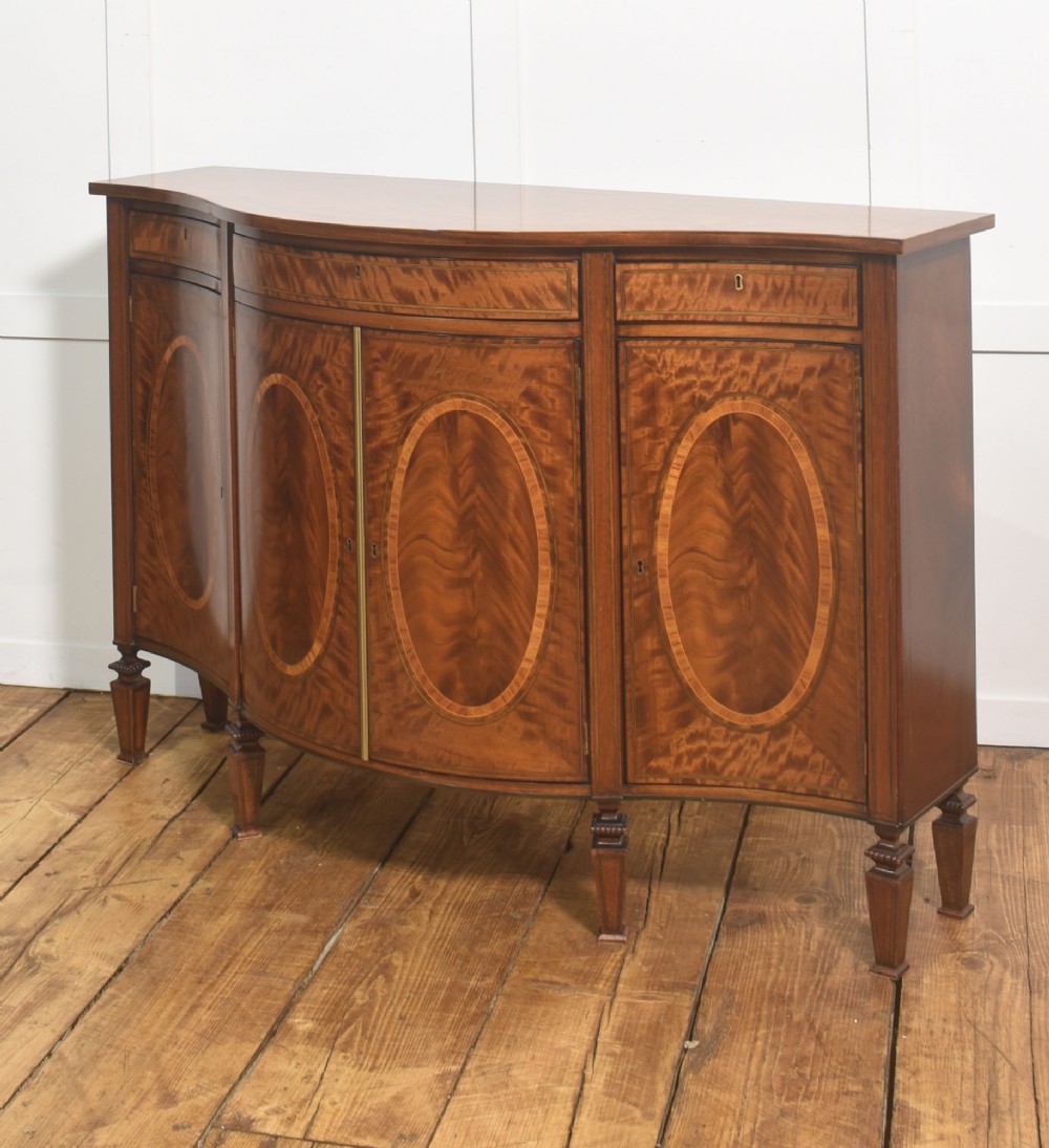 A Sheraton Revival bow-front wooden sideboard featuring oval inlaid veneer door panels, top drawers, and square tapered legs. View X