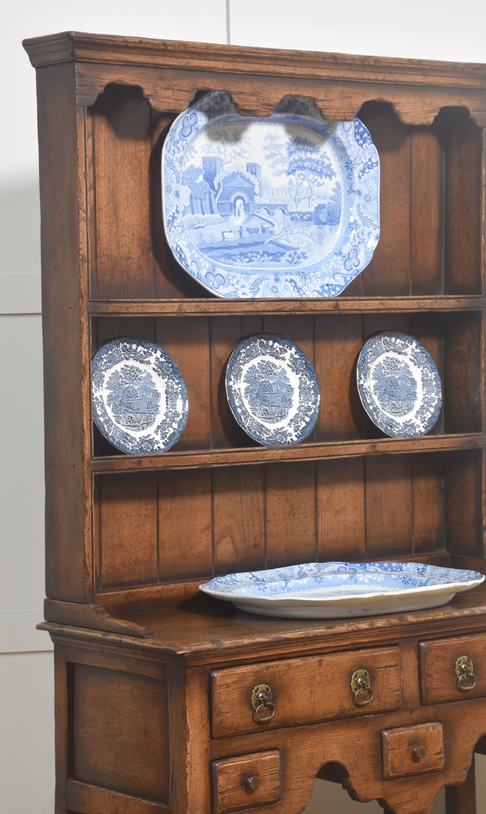 An antique warm brown oak dresser featuring an upper three-shelf plate rack with a scalloped canopy, sitting above five drawers and an open pot shelf with a turned central support. View V