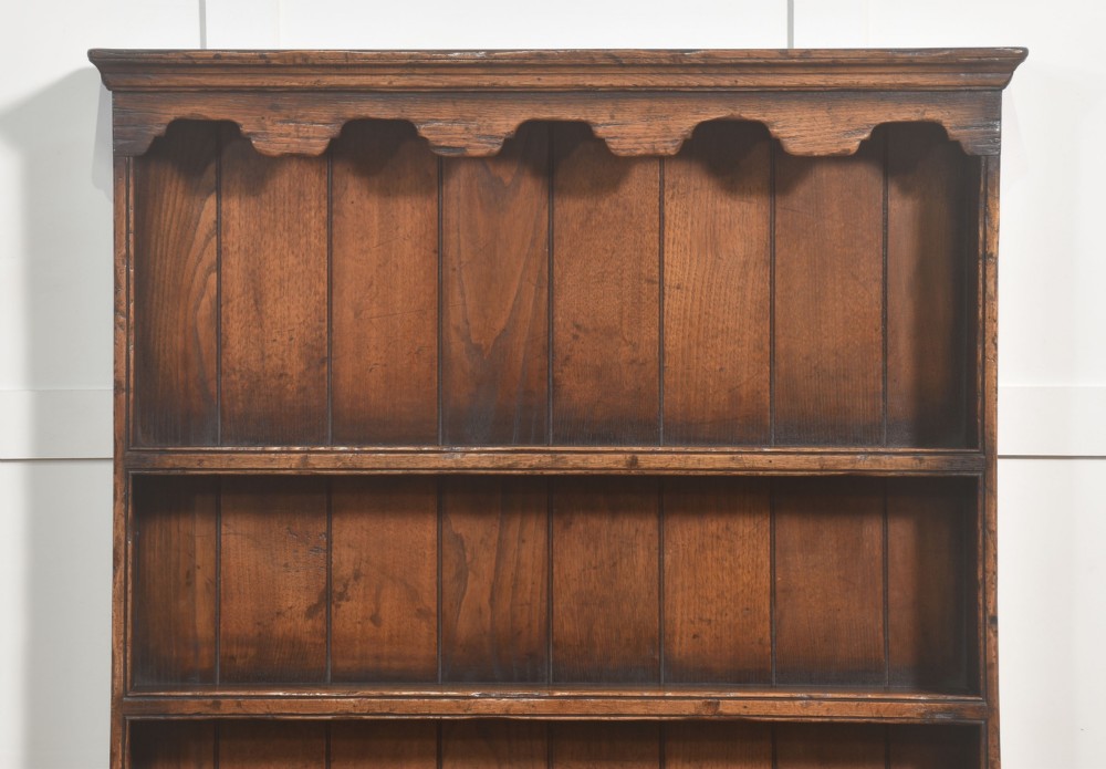 An antique warm brown oak dresser featuring an upper three-shelf plate rack with a scalloped canopy, sitting above five drawers and an open pot shelf with a turned central support. View VIII