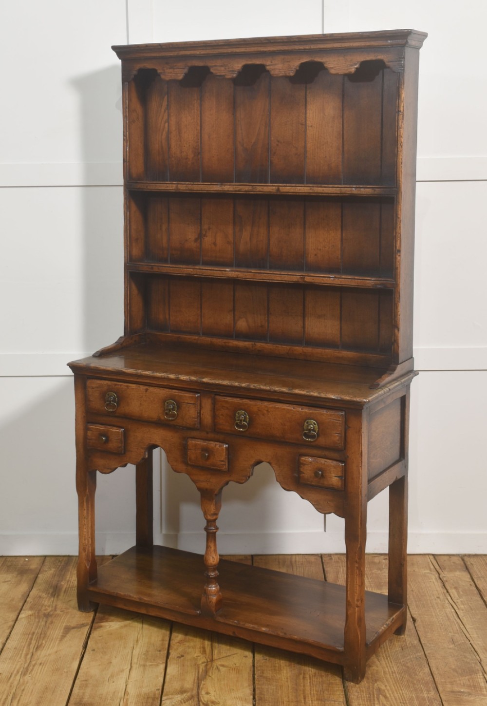 An antique warm brown oak dresser featuring an upper three-shelf plate rack with a scalloped canopy, sitting above five drawers and an open pot shelf with a turned central support. View X