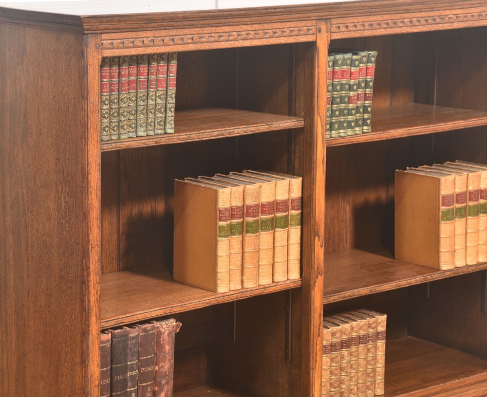 A low freestanding solid oak open bookcase featuring a double bay configuration, carved frieze detail, and adjustable shelves housing books. View VI