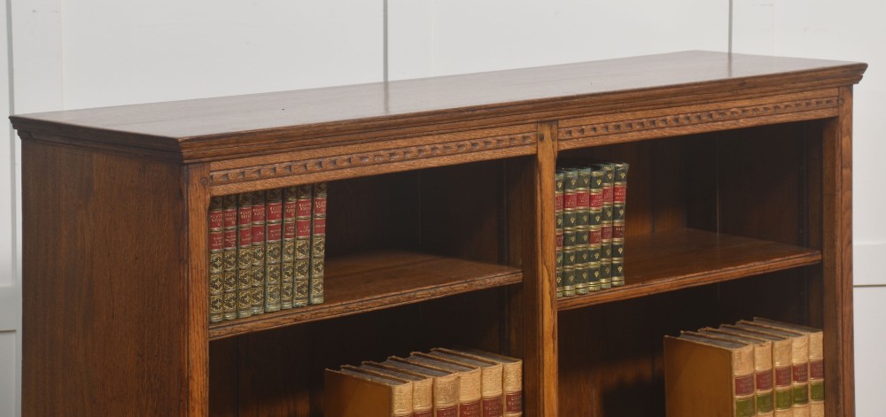 A low freestanding solid oak open bookcase featuring a double bay configuration, carved frieze detail, and adjustable shelves housing books. View VII