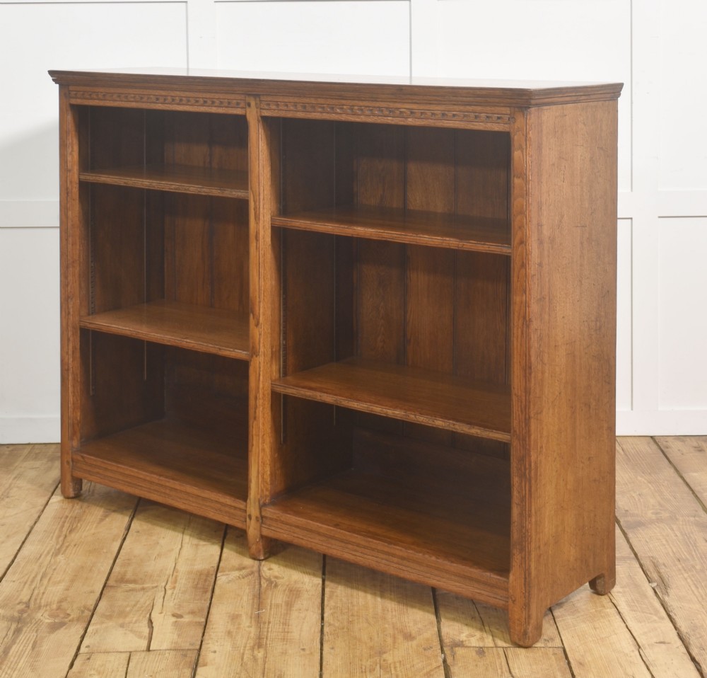 A low freestanding solid oak open bookcase featuring a double bay configuration, carved frieze detail, and adjustable shelves housing books. View IX
