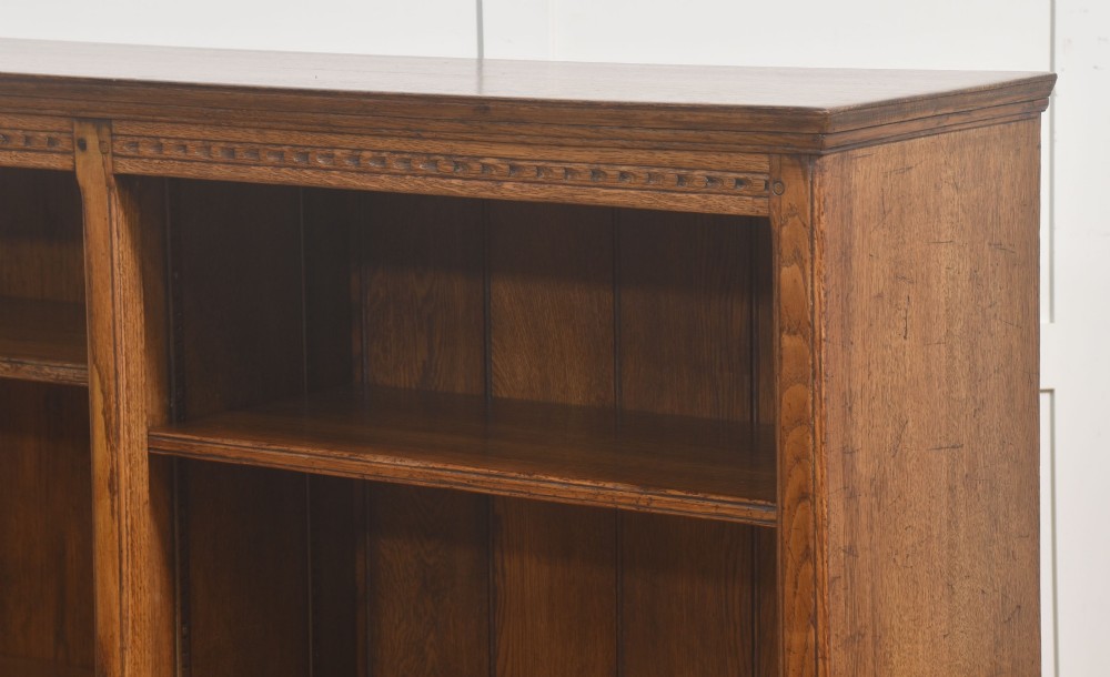 A low freestanding solid oak open bookcase featuring a double bay configuration, carved frieze detail, and adjustable shelves housing books. View X