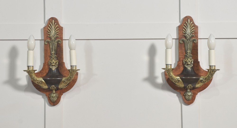 A pair of Empire style wall lights featuring shaped burr walnut backplates decorated with brass lion masks, anthemion crests, and twin scrolling candle arms.