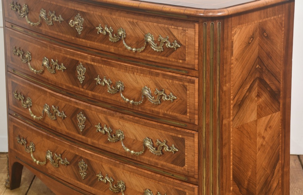 A French Louis XV style serpentine chest of drawers featuring geometric wood marquetry and decorative brass handles. View X