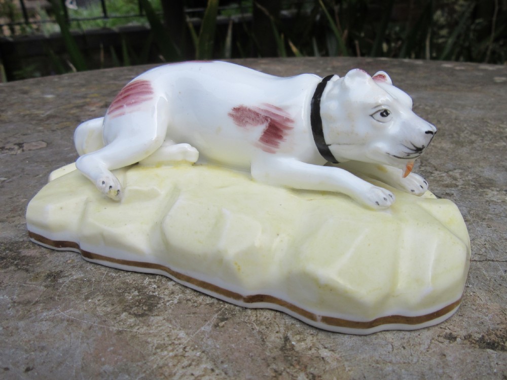 An antique Victorian white porcelain figurine of a reclining mastiff dog with reddish patches on a pale yellow gilded base.