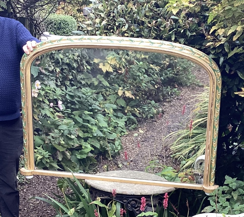 An arched Victorian giltwood overmantel mirror featuring hand-painted lily of the valley floral motifs along the upper frame and white ceramic feet.