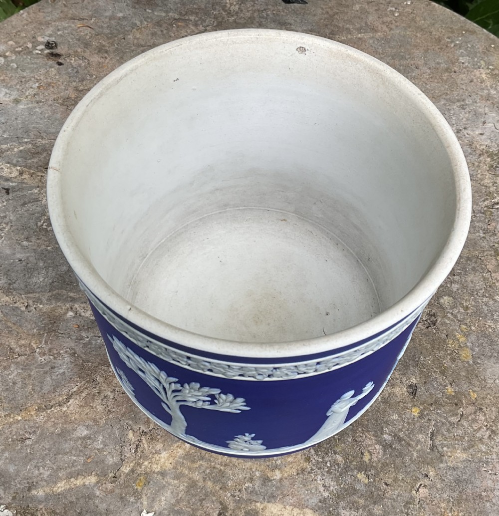 A cobalt blue Wedgwood Jasperware cylindrical jardiniere decorated with white classical relief figures, trees, a top floral garland border, resting on three white feet. View IV
