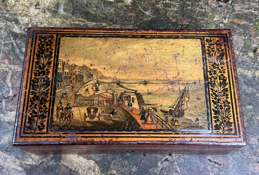 An antique rectangular wooden box with black penwork illustration of Brighton Chain Pier framed by detailed foliate borders.