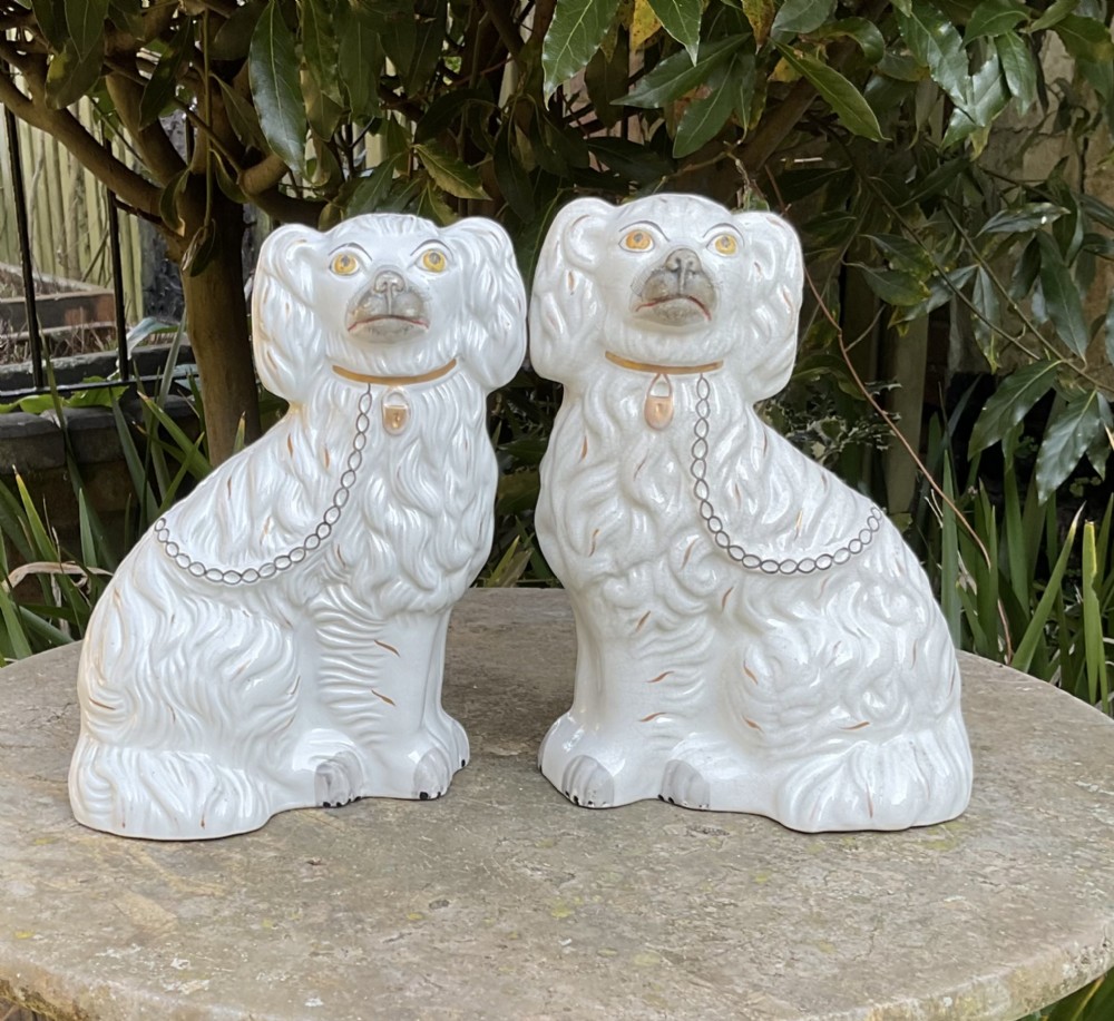 A pair of antique Victorian white earthenware Staffordshire spaniel dog figurines featuring gilded collars, chains, and painted facial features.