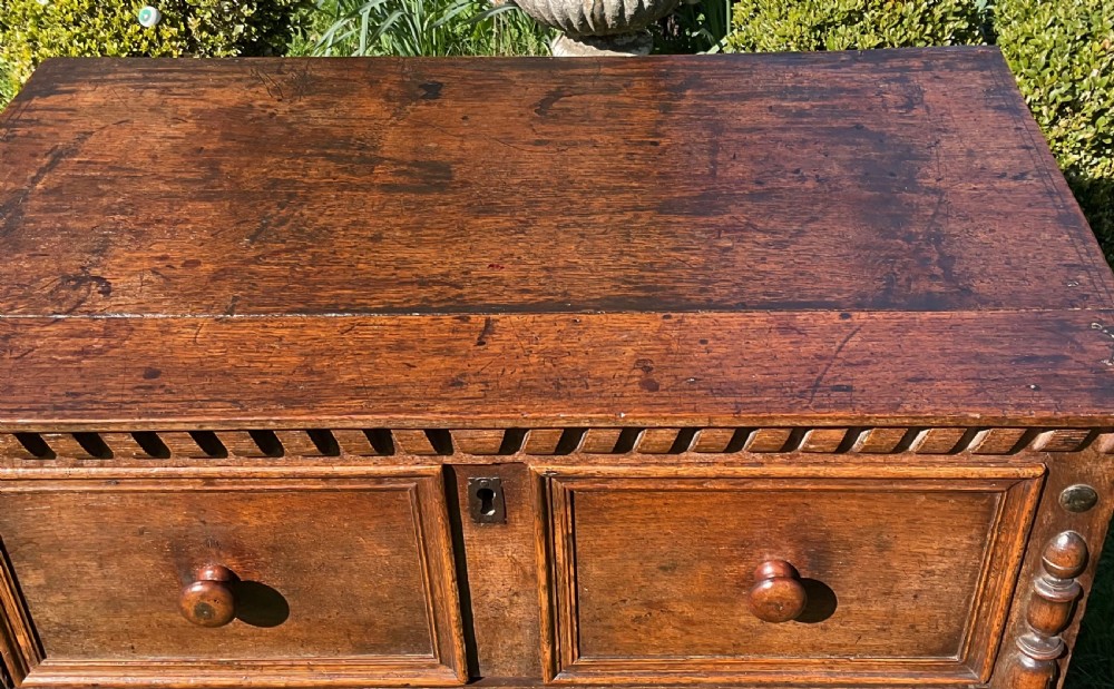 A 17th-century English dark oak chest of drawers featuring geometric panel mouldings, turned wooden knobs, split-spindle side mouldings, and a scalloped base. View V