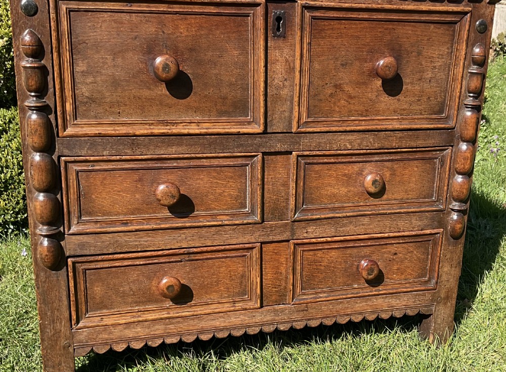 A 17th-century English dark oak chest of drawers featuring geometric panel mouldings, turned wooden knobs, split-spindle side mouldings, and a scalloped base. View VII