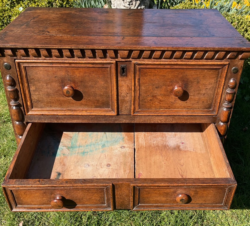 A 17th-century English dark oak chest of drawers featuring geometric panel mouldings, turned wooden knobs, split-spindle side mouldings, and a scalloped base. View XII
