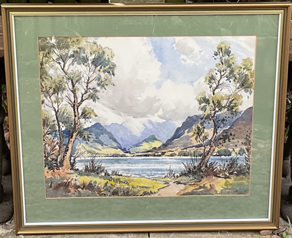 A framed watercolour landscape painting depicting a mountain lake framed by trees under a cloudy sky, mounted in green and framed in gold.