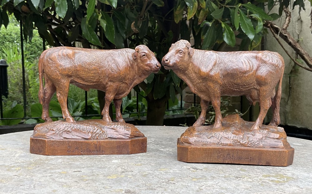 A pair of hand-carved wooden sculptures depicting a bull and a cow standing on naturalistic carved bases with artist signatures.