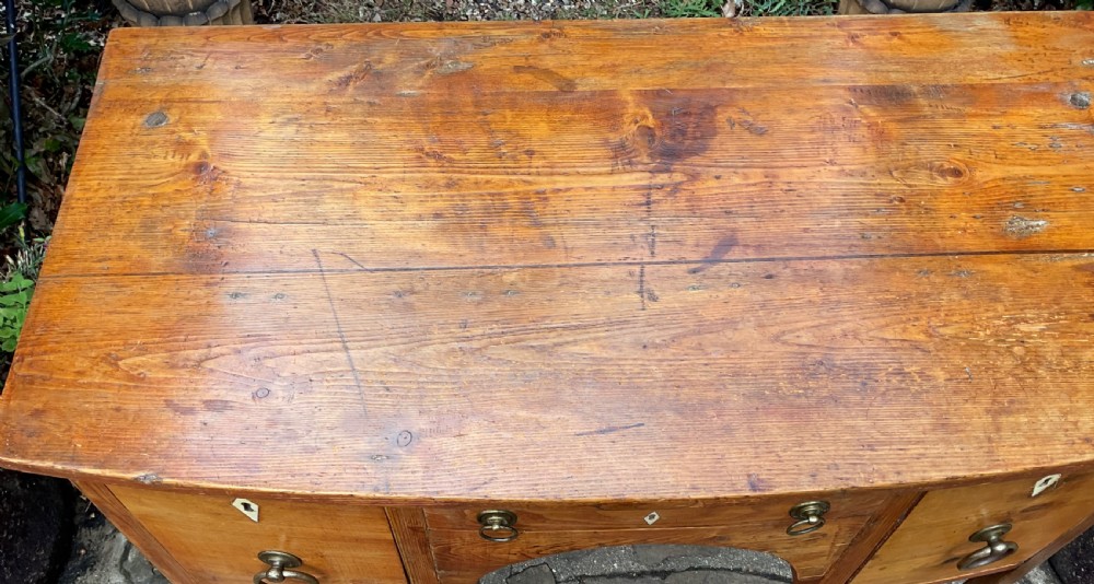 A Georgian pine bowfront sideboard featuring a arched central apron with a shallow drawer, flanked by two side drawers, raised on four tapered square legs. View VII