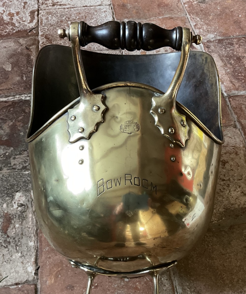 A Victorian brass coal helmet scuttle resting on a pedestal base, complete with an ebonised wooden handle and a matching brass scoop. View IX