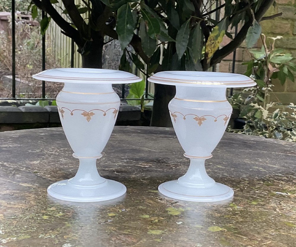 A pair of translucent white opaline glass urn-shaped vases featuring wide flared rims, turned pedestal stems, and delicate hand-painted gold leaf floral swags.