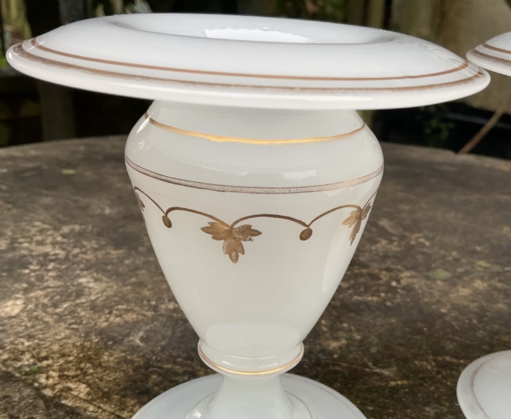 A pair of translucent white opaline glass urn-shaped vases featuring wide flared rims, turned pedestal stems, and delicate hand-painted gold leaf floral swags. View V