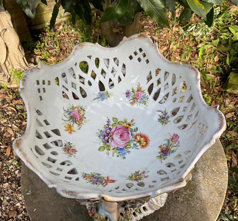 An ornate German porcelain centrepiece featuring a pierced lattice bowl encrusted with colourful three-dimensional flowers, supported by a central pedestal with a seated winged cherub on a scrollwork base. View V