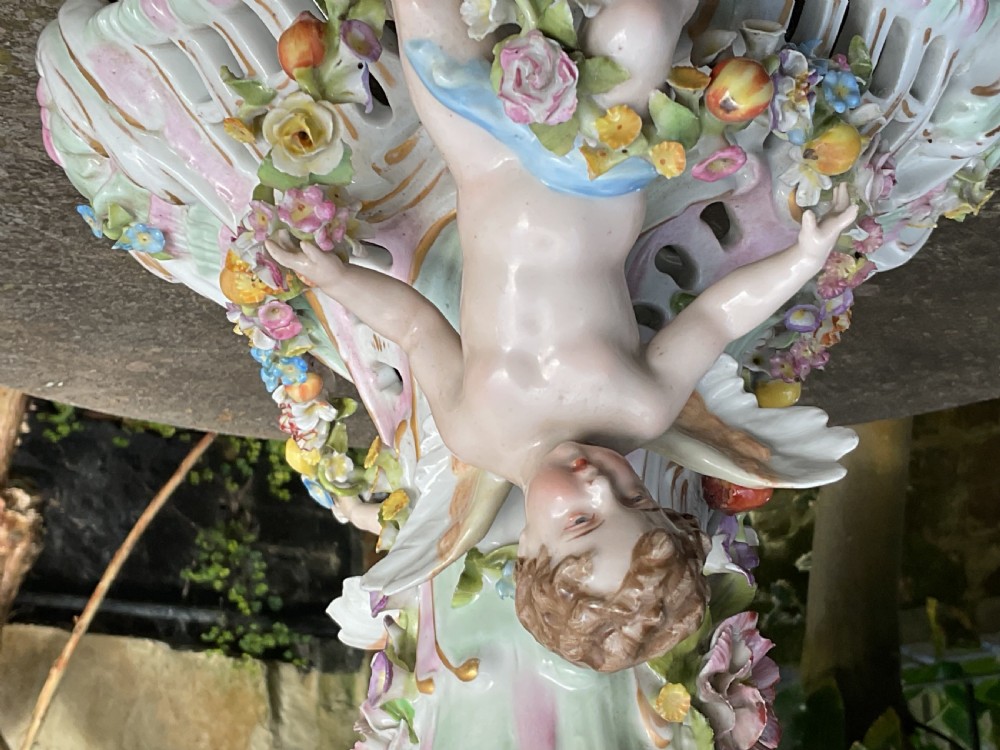 An ornate German porcelain centrepiece featuring a pierced lattice bowl encrusted with colourful three-dimensional flowers, supported by a central pedestal with a seated winged cherub on a scrollwork base. View XII