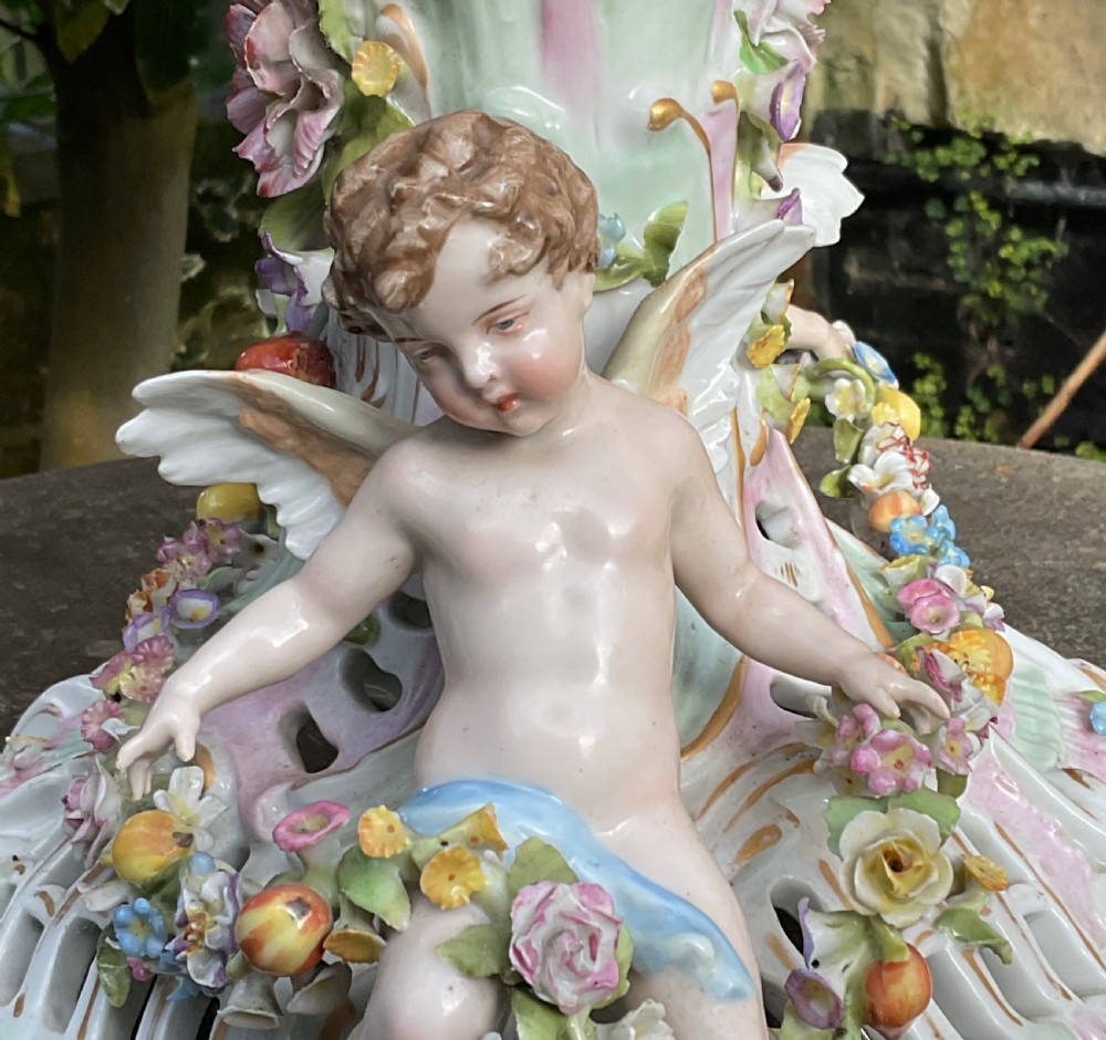 An ornate German porcelain centrepiece featuring a pierced lattice bowl encrusted with colourful three-dimensional flowers, supported by a central pedestal with a seated winged cherub on a scrollwork base. View XV