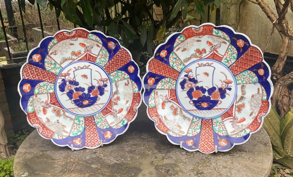 A pair of late nineteenth-century Japanese Imari porcelain plates featuring scalloped rims, painted in cobalt blue, rust red, and green with central floral basket motifs and patterned panel borders.