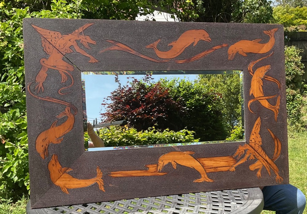 A large rectangular Arts and Crafts style wall mirror featuring a bevelled glass plate surrounded by a textured frame decorated with mythical dragons and dolphins.