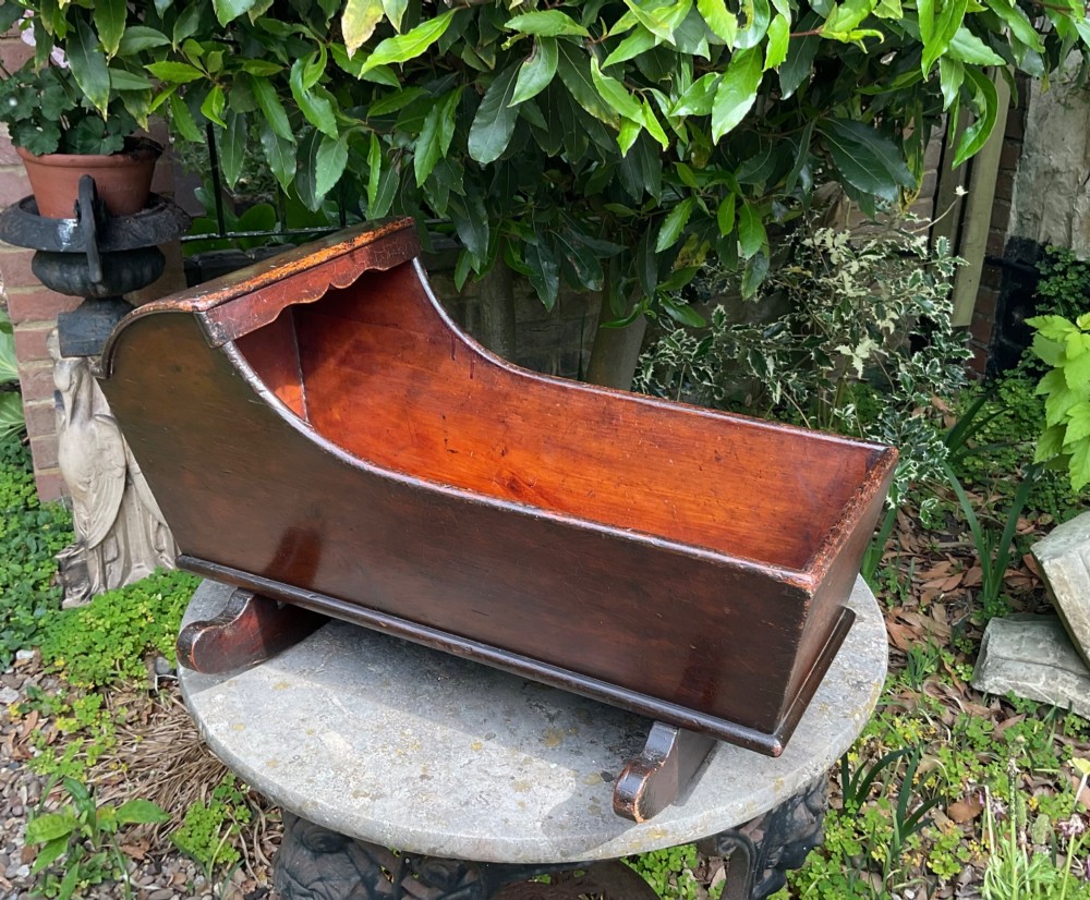 A Georgian pine miniature doll cradle featuring a hooded canopy, scalloped front moulding, and curved wooden rockers in a warm brown finish. View II