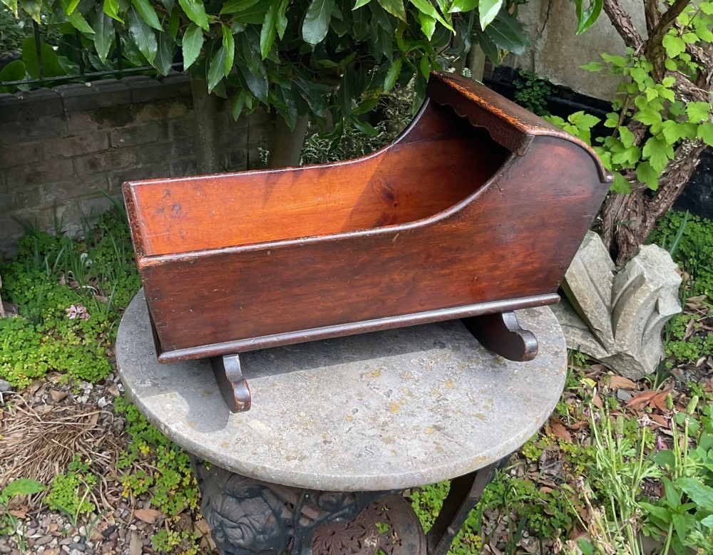 A Georgian pine miniature doll cradle featuring a hooded canopy, scalloped front moulding, and curved wooden rockers in a warm brown finish. View V