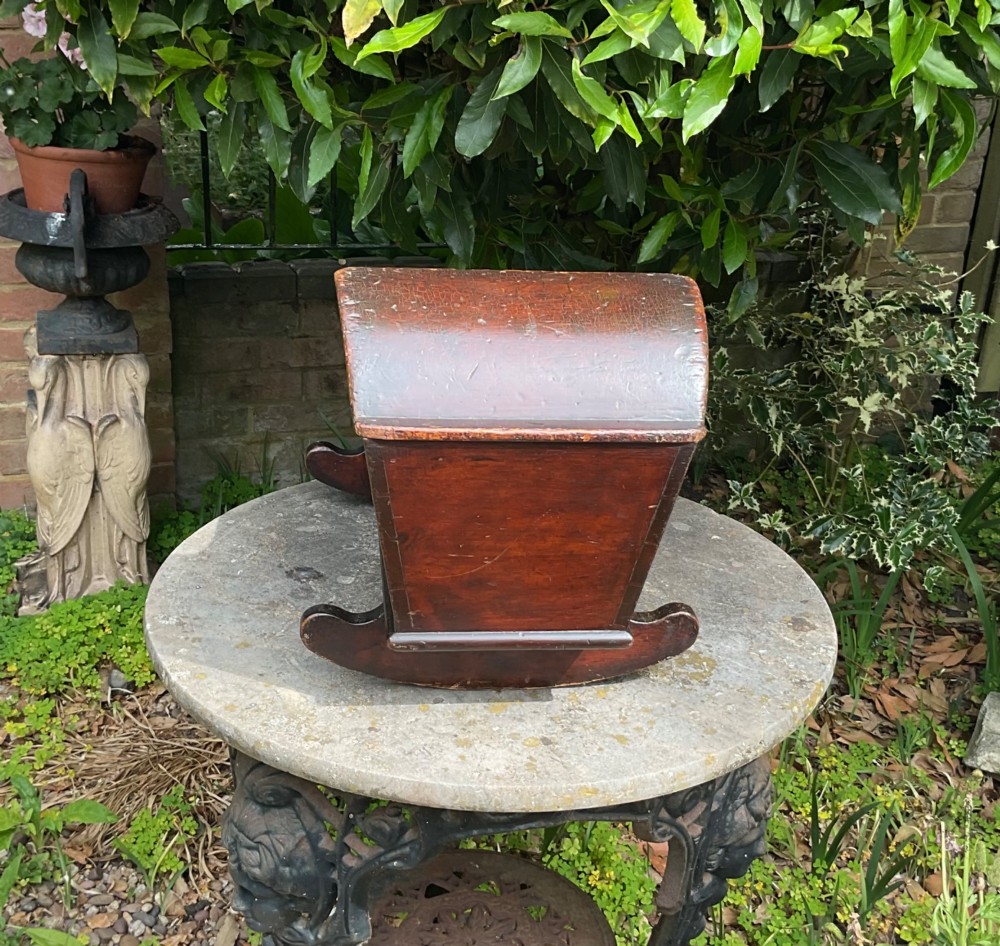 A Georgian pine miniature doll cradle featuring a hooded canopy, scalloped front moulding, and curved wooden rockers in a warm brown finish. View VI