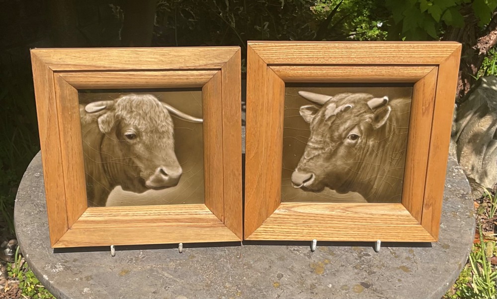 A pair of square sepia-toned glazed ceramic tiles featuring relief portraits of a bull and a cow, each enclosed within a wooden oak frame.