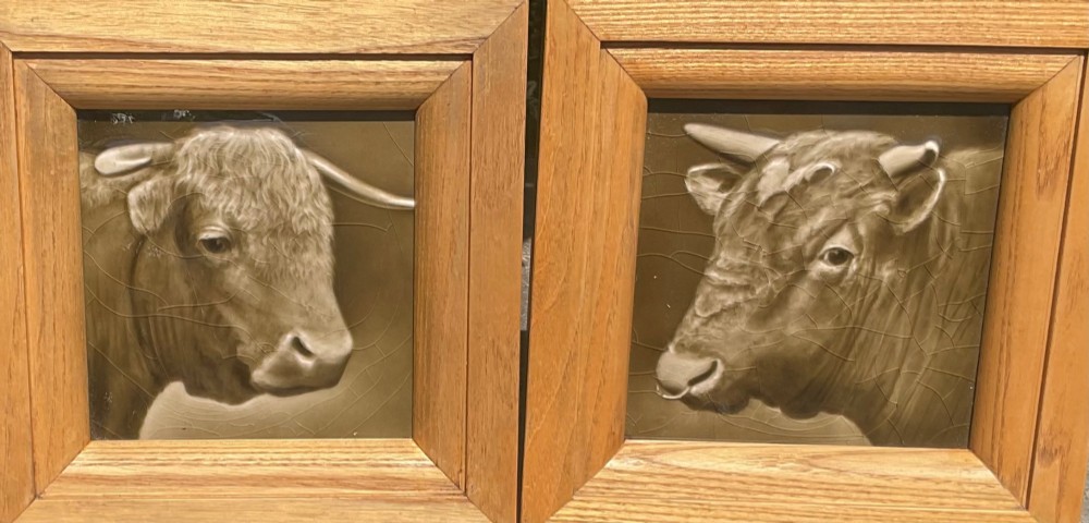 A pair of square sepia-toned glazed ceramic tiles featuring relief portraits of a bull and a cow, each enclosed within a wooden oak frame. View II