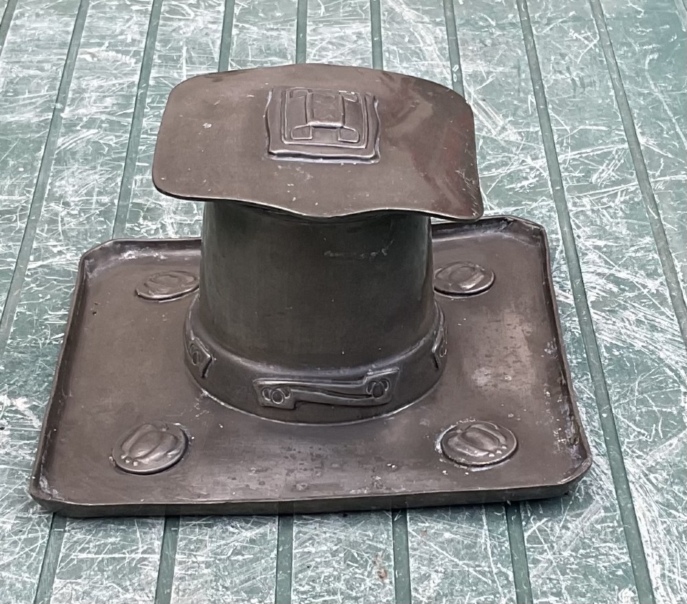 An Arts and Crafts Tudric pewter inkwell featuring a square base tray with raised relief motifs and a central cylindrical body topped with a square lid.