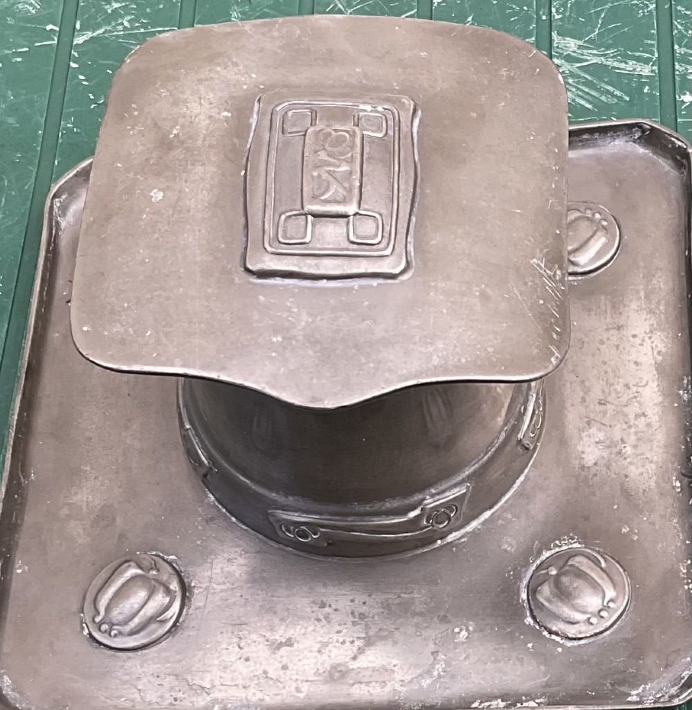 An Arts and Crafts Tudric pewter inkwell featuring a square base tray with raised relief motifs and a central cylindrical body topped with a square lid. View II