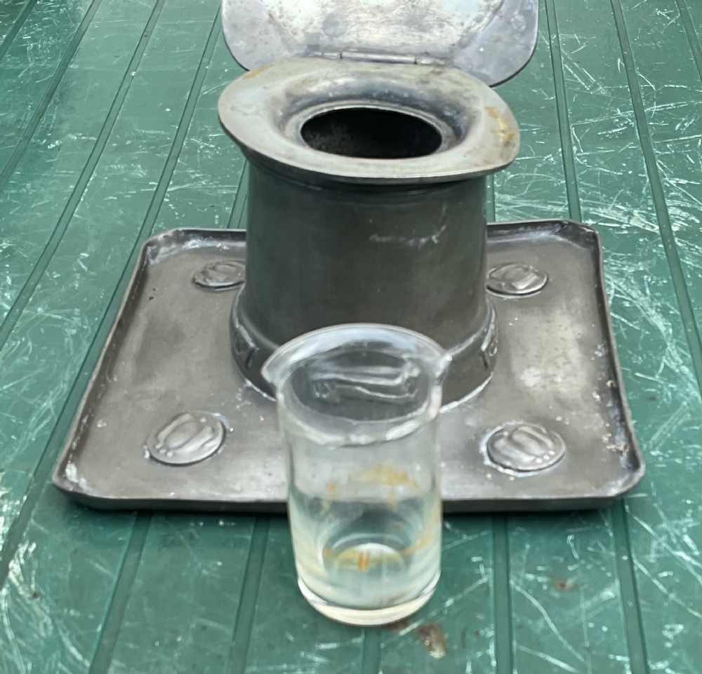 An Arts and Crafts Tudric pewter inkwell featuring a square base tray with raised relief motifs and a central cylindrical body topped with a square lid. View IV