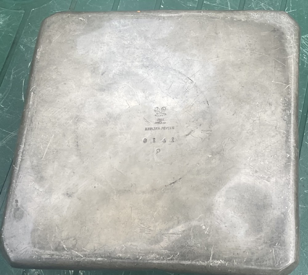 An Arts and Crafts Tudric pewter inkwell featuring a square base tray with raised relief motifs and a central cylindrical body topped with a square lid. View V