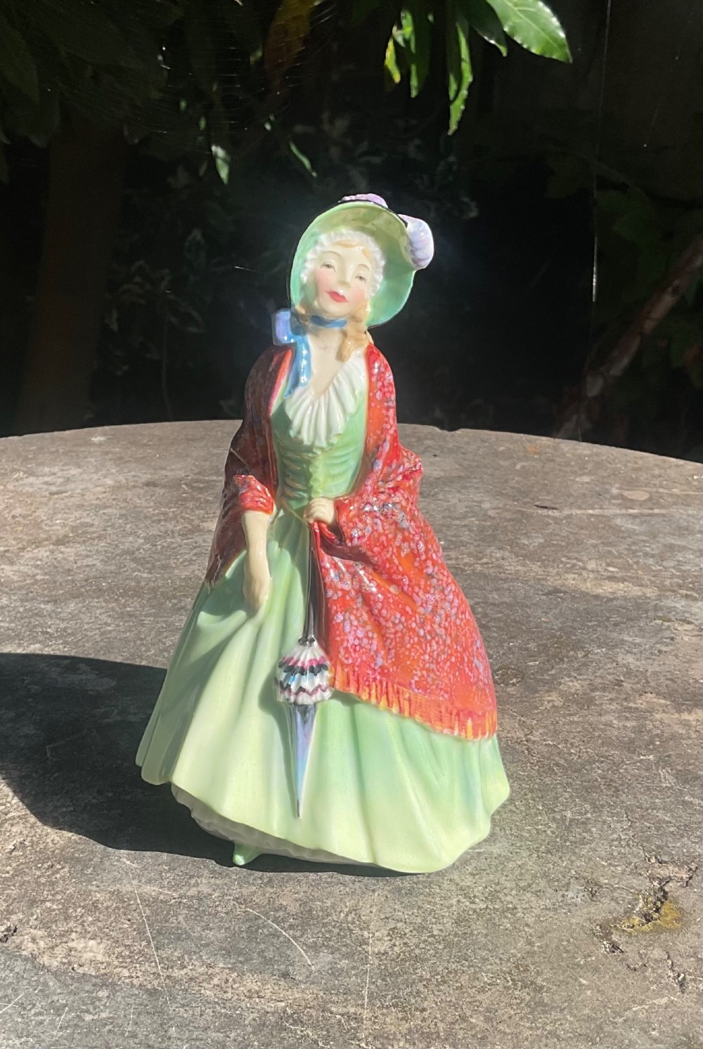 A Royal Doulton fine bone china figurine depicting a woman in a green gown and bonnet holding a parasol and draped in a patterned red shawl.