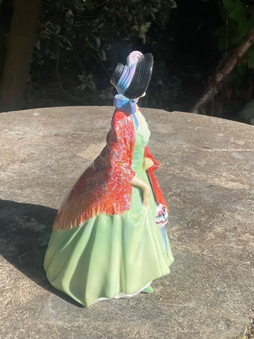 A Royal Doulton fine bone china figurine depicting a woman in a green gown and bonnet holding a parasol and draped in a patterned red shawl. View IV