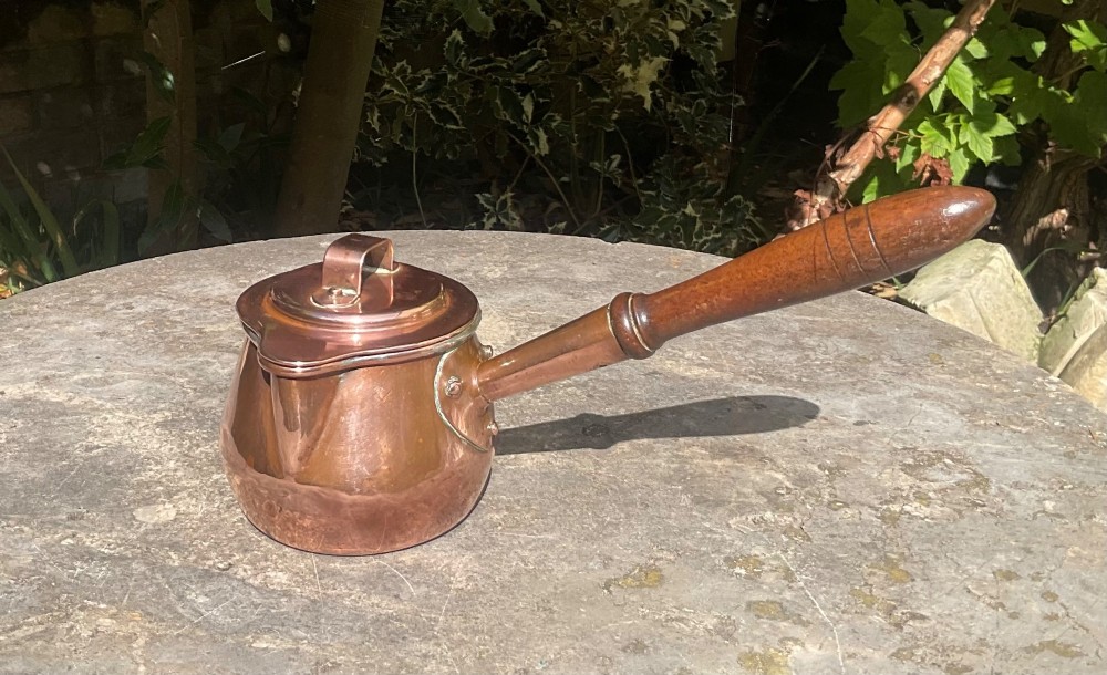 An 1830s Georgian copper brandy warmer featuring a polished copper vessel, fitted lid with loop handle, pouring spout, and an extended turned wooden handle.