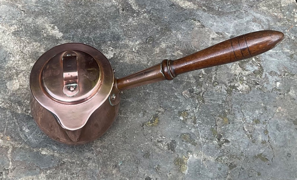 An 1830s Georgian copper brandy warmer featuring a polished copper vessel, fitted lid with loop handle, pouring spout, and an extended turned wooden handle. View II