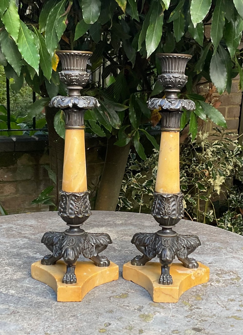 A pair of Regency period candlesticks with yellow marble column stems, classical dark bronze sconces, acanthus collars, and bronze paw feet on triform marble bases.